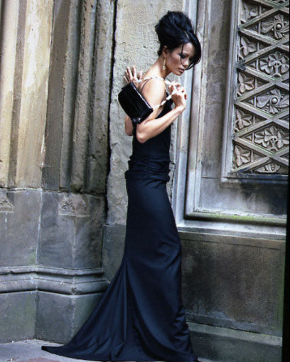 Woman in a black evening gown standing against a stone wall. holding a Darby Scott Handbag