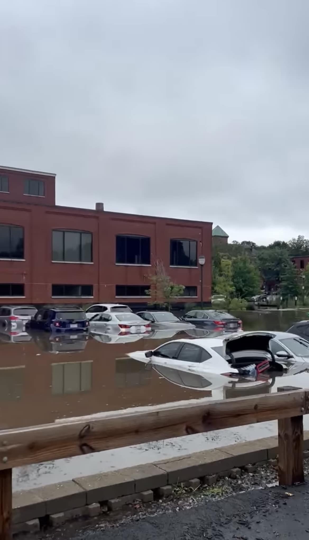 Flooded parking lot with cars partially submerged, red brick building in the background. - Mills in North Andover