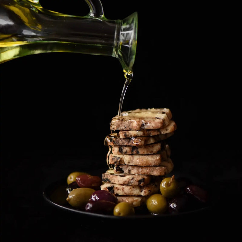 Bottle of olive oil being poured over dish of savory olive biscuits and olives