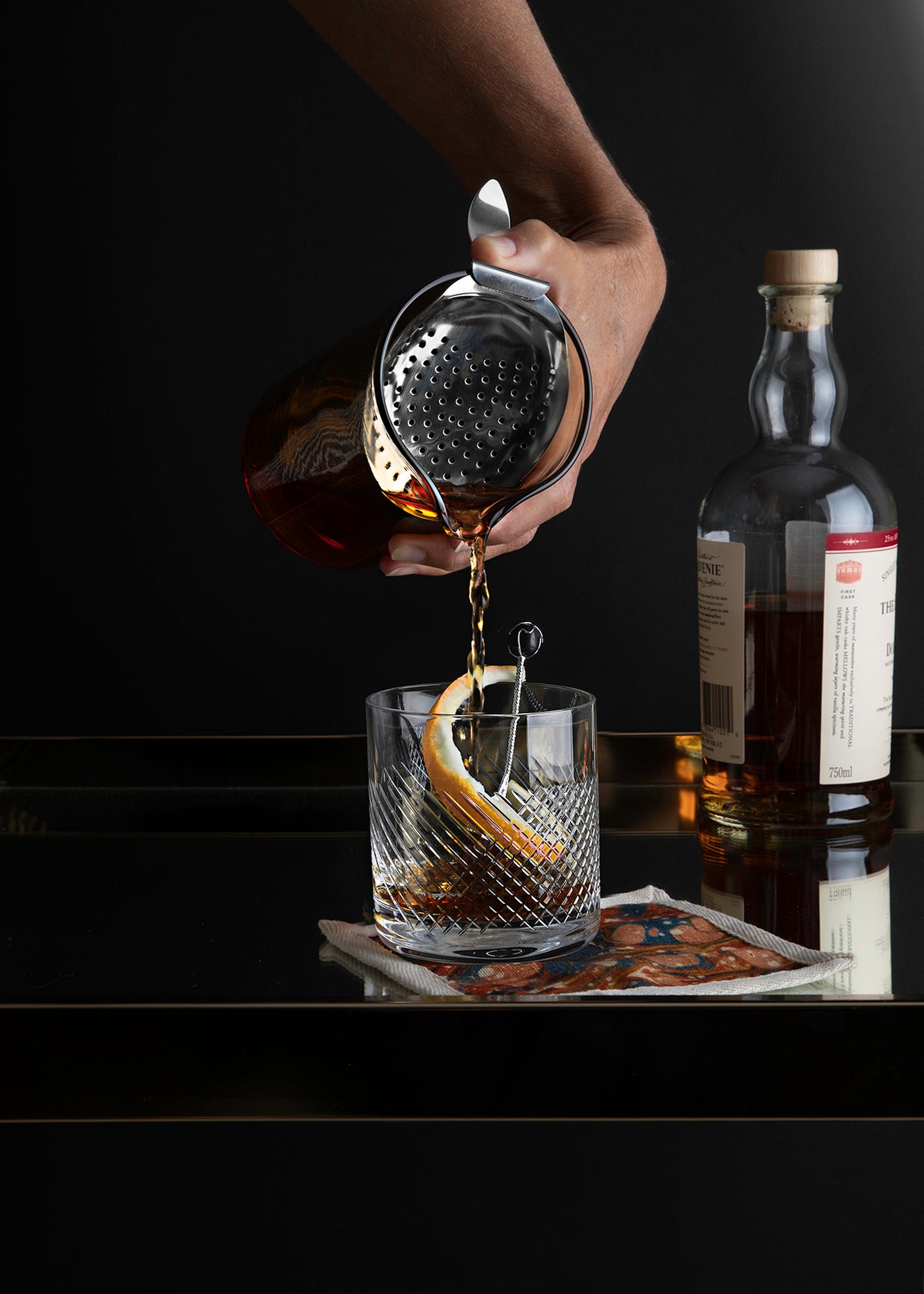 Person using a mixing glass with thumb-held strainer to pour a drink into cut-crystal glass