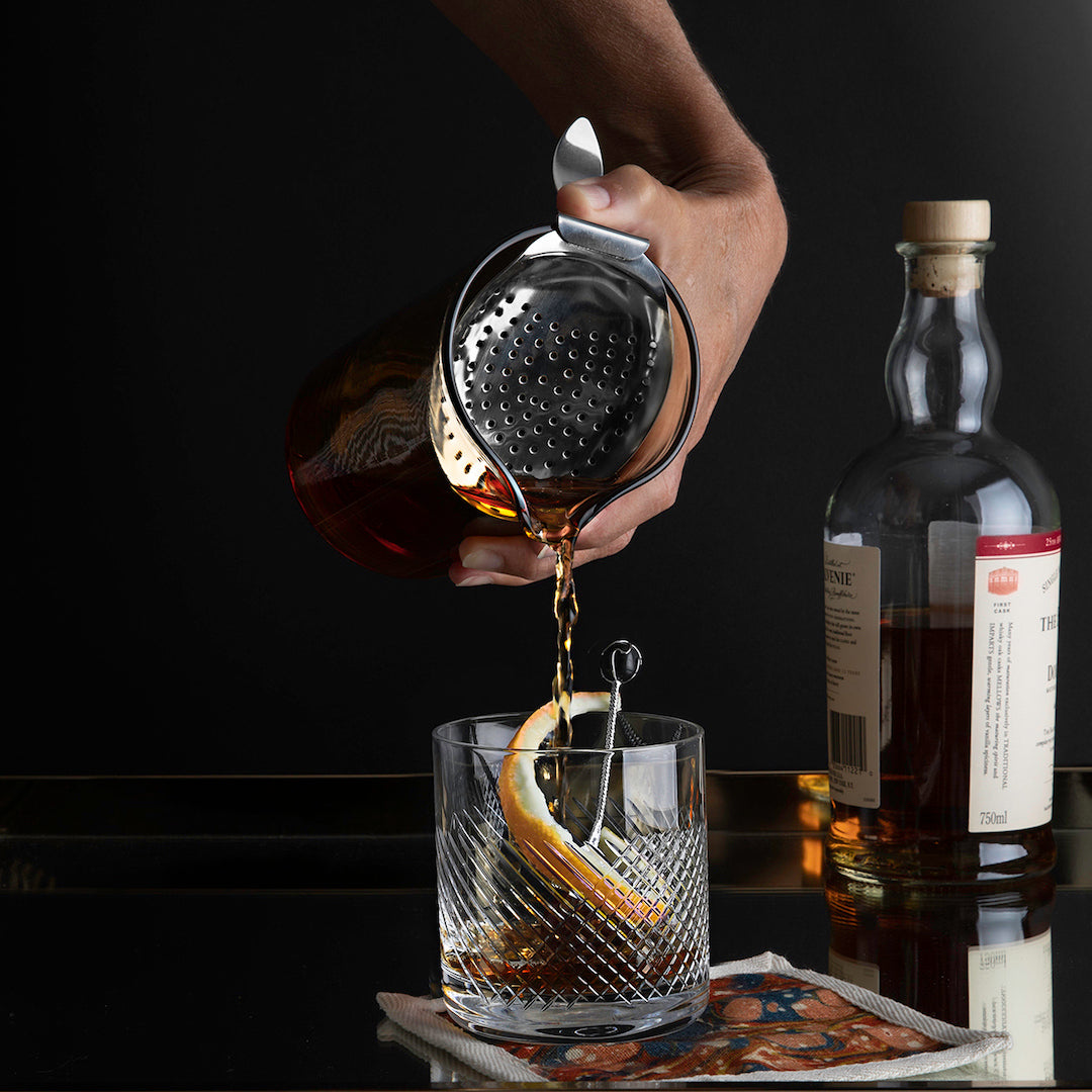 Person using a mixing glass with thumb-held strainer to pour a drink into cut-crystal glass