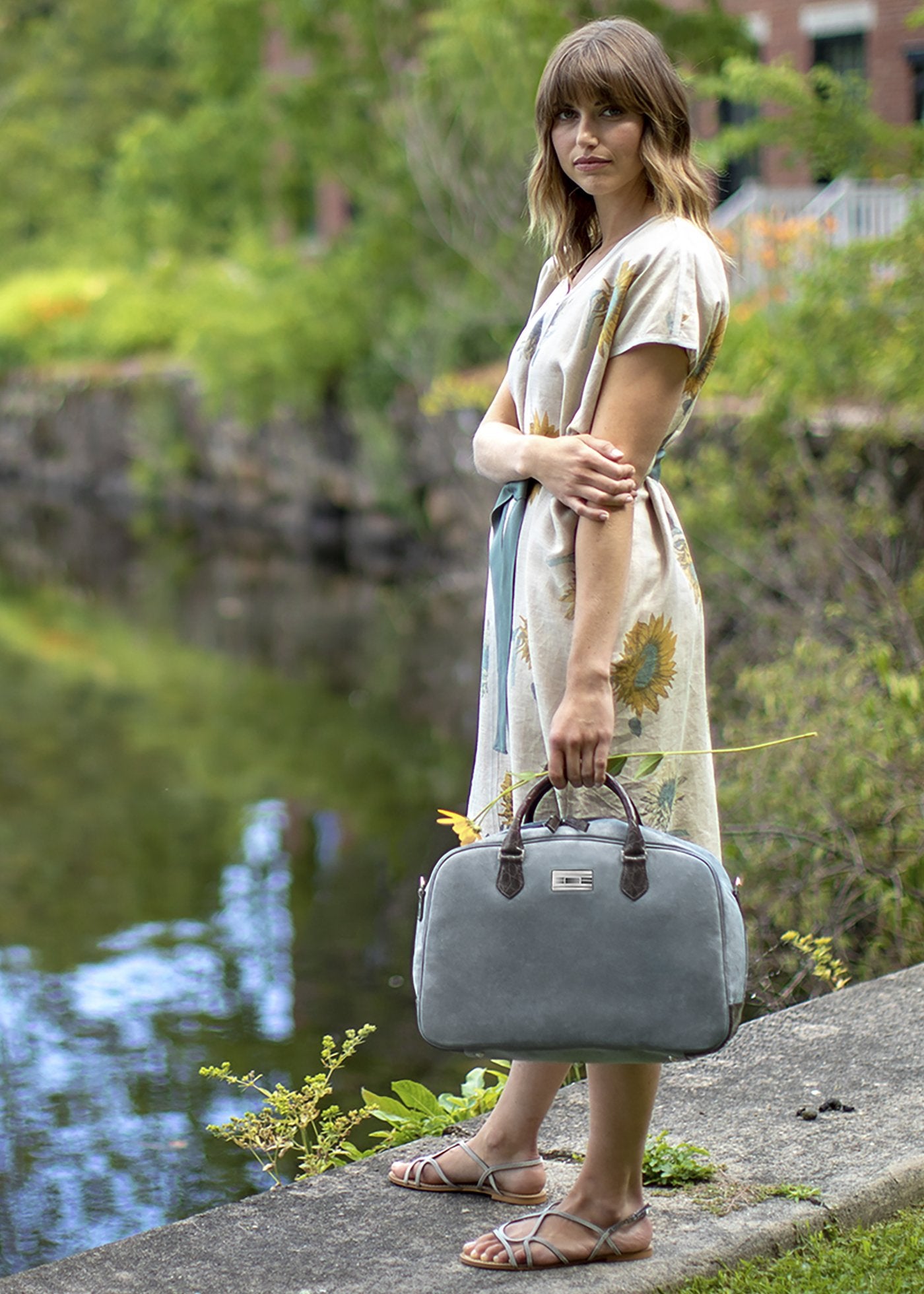 Model outdoors holding Newport Travel Satchel Bag in Denim Blue Suede with Brown Croc Trim - Darby Scott